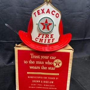 May include: A red plastic fire chief helmet with a white front shield featuring the Texaco logo and text. It sits atop a square cardboard box printed with the text: Trust your car to the man who wears the star, manufactured for Texaco by Brown & Bigelow.