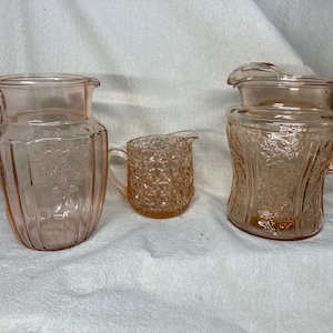 May include: Three vintage pink glass pitchers with embossed floral designs. The pitchers are in various sizes and have a delicate, feminine aesthetic.