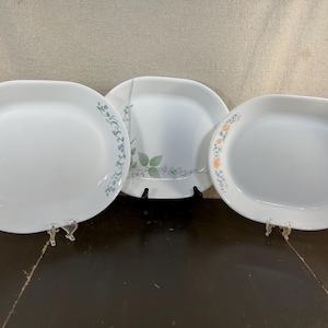 以下が含まれることがあります： 花柄の白い楕円形と正方形のプレート3枚。 プレートは白い背景に青、緑、オレンジの花柄があります。 プレートは透明なスタンドに展示されています。