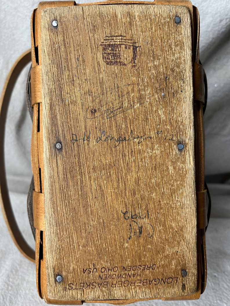May include: A Longaberger Basket with a brown leather handle and a wooden base. The basket is stamped with the words "Longaberger Baskets, Handwoven, Dresden, Ohio, USA".
