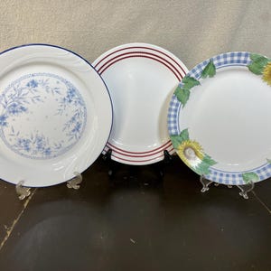 May include: Three decorative plates with different designs. One plate has a blue floral pattern, another has red stripes, and the third features sunflowers and a blue gingham border. Each plate is white.