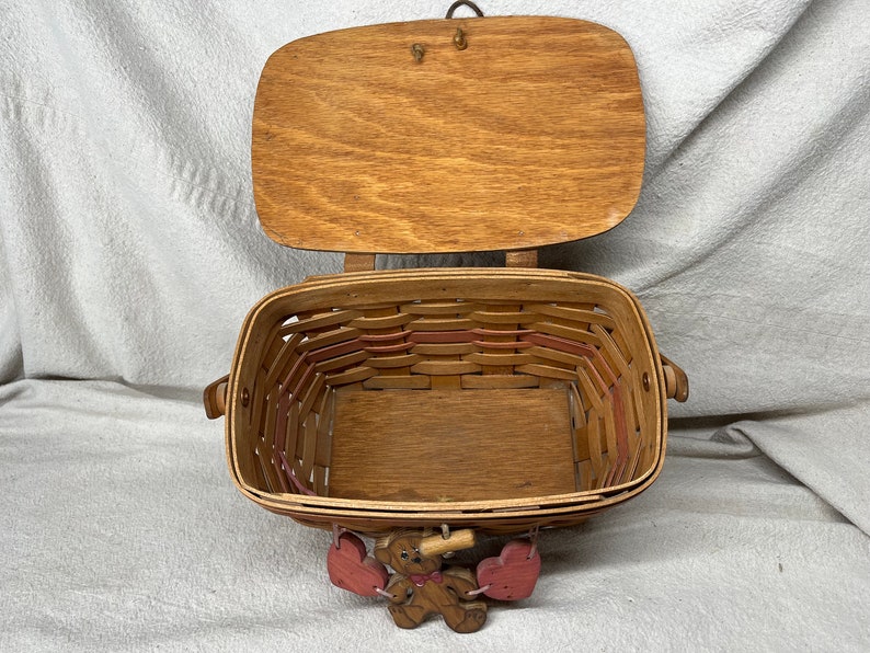 May include: A wooden basket with a hinged lid and a woven design. The basket is decorated with pink hearts and a small wooden bear figurine.