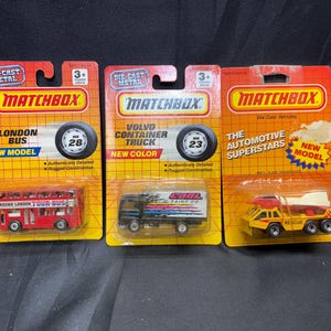 May include: Three Matchbox die-cast metal toy vehicles on their original packaging. The collection includes a red London Bus, a Volvo Container Truck, and a yellow rescue truck. Each package features the Matchbox logo and vehicle details.
