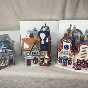 May include: Three miniature Christmas village buildings, each with a unique design and snowy roofs. The buildings are made of ceramic and feature intricate details, including windows, doors, and signs. The building on the left is a red and white building with the text "Town Tavern" on the sign. The building in the center is a brown and white building with the text "Terry's Treats" on the sign. The building on the right is a blue and red building with the text "Christmas Bread Bakers" on the sign.