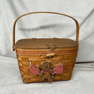 May include: A small wooden basket with a brown lid and a woven base. The basket has a brown teddy bear with pink hearts hanging from it. The basket has a wooden handle.