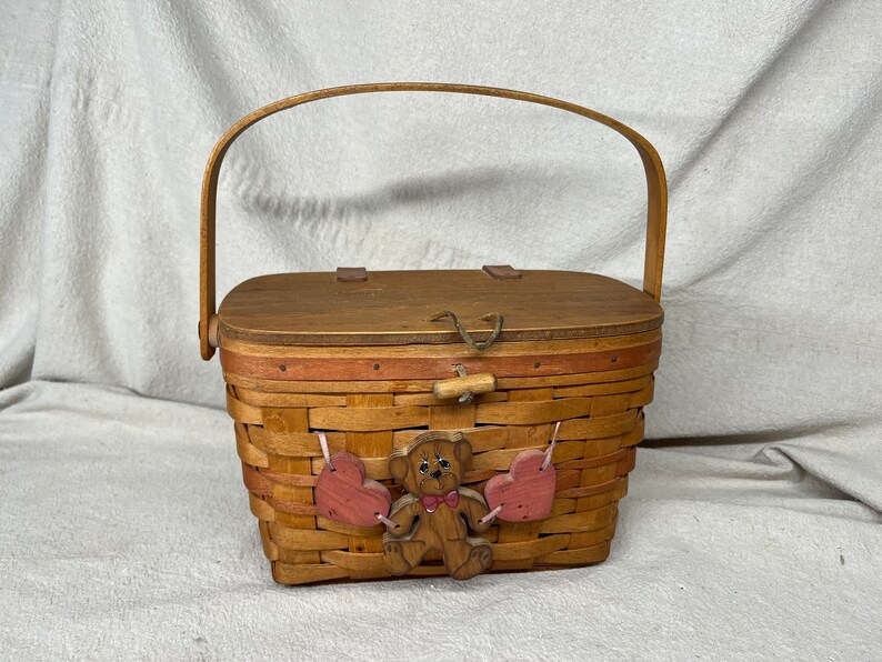 May include: A small wooden basket with a brown lid and a woven base. The basket has a brown teddy bear with pink hearts hanging from it. The basket has a wooden handle.