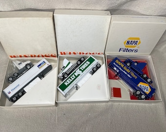 Puede incluir: Tres camiones de juguete en miniatura en cajas. Los camiones son blancos, verdes y azules. Las cajas son blancas con la palabra "WINROSS" impresa en ellas. Los camiones están etiquetados con los nombres de las empresas: "Chevron", "Kelly Tires" y "NAPA Gold Filters".