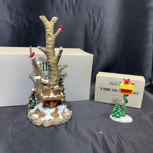 Puede incluir: Una escena invernal decorativa con un abedul con pájaros rojos y una casita para pájaros, y una casita para pájaros separada en un poste. La escena está ambientada sobre una base nevada con pequeños árboles verdes. El texto en la caja dice "A Home for the Holidays".