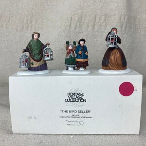 May include: A set of three hand-painted porcelain figurines depicting a bird seller with two children. The seller is holding two birdcages, one in each hand. The children are holding birdcages. The figurines are part of the Heritage Village Collection and are titled "The Bird Seller".
