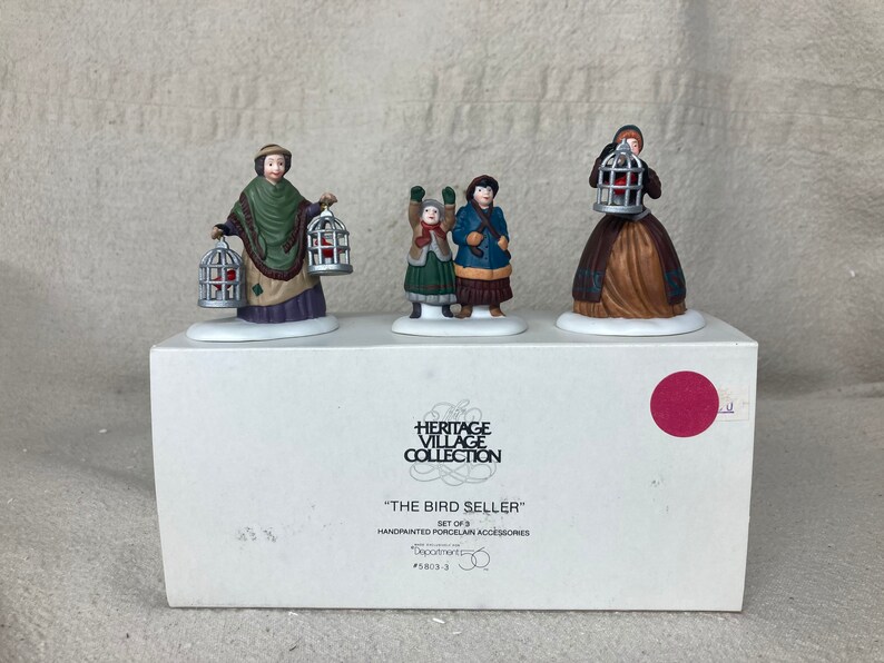 May include: A set of three hand-painted porcelain figurines depicting a bird seller with two children. The seller is holding two birdcages, one in each hand. The children are holding birdcages. The figurines are part of the Heritage Village Collection and are titled "The Bird Seller".