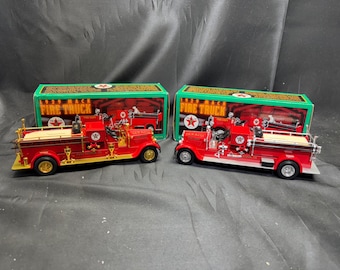 May include: Two red 1929 Mack Fire Truck model toys with gold and silver accents. Each toy is displayed in front of a green box with the words "1929 Mack Fire Truck" printed on it. The toys have black tires.