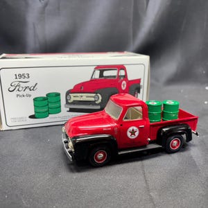 May include: A red 1953 Ford Pick-Up truck model with black accents and a white box. The truck has a Texaco logo and green barrels in the bed. The box shows a similar truck and the words "1953 Ford Pick-Up".