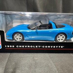Puede incluir: Un coche modelo azul de un Chevrolet Corvette de 1998 en una vitrina transparente. El coche tiene ruedas plateadas y un interior negro. La caja tiene el texto "1998 CHEVROLET CORVETTE" y el logotipo de Chevrolet.