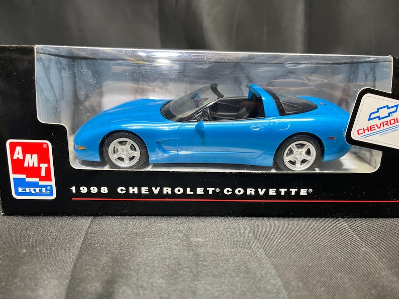 Puede incluir: Un coche modelo azul de un Chevrolet Corvette de 1998 en una vitrina transparente. El coche tiene ruedas plateadas y un interior negro. La caja tiene el texto "1998 CHEVROLET CORVETTE" y el logotipo de Chevrolet.