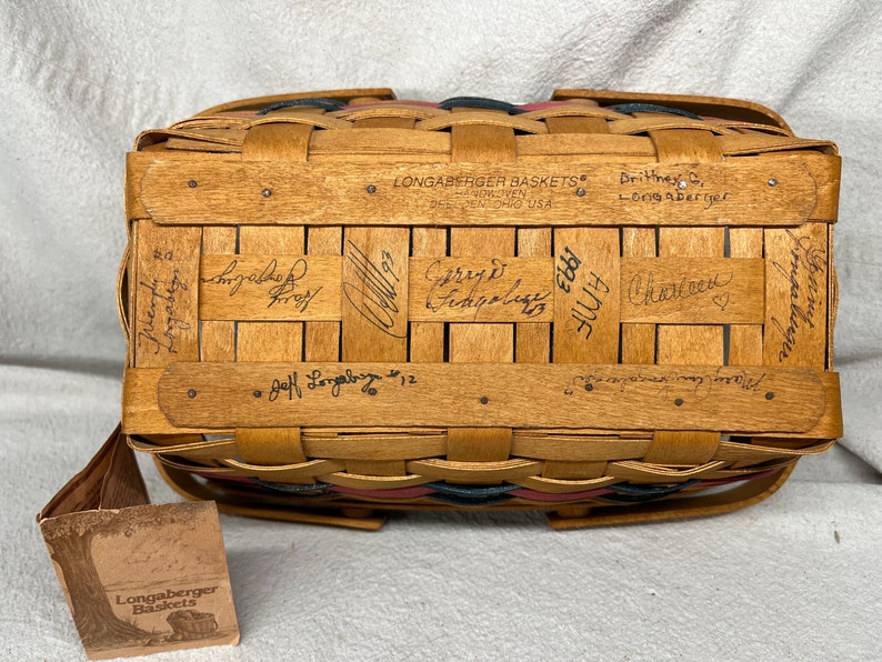 May include: A Longaberger basket with a red, white, and blue woven design. The basket is signed by Jeff Longaberger and several other people. The basket has a tag that says "Longaberger Baskets".