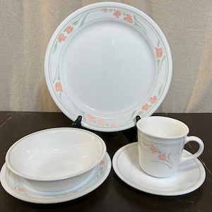 May include: A set of white ceramic dinnerware with a floral design in shades of pink and green. The set includes a dinner plate, a bowl, a cup, and a saucer.