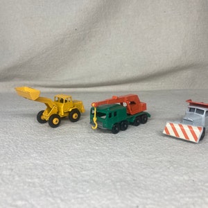 May include: Three toy construction vehicles: a yellow front loader, a green crane truck with an orange boom, and a gray dump truck with an orange bed and a red and white snow plow.