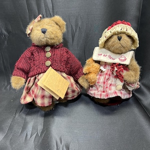 May include: Two plush teddy bears. One bear wears a burgundy sweater and plaid skirt, with a pink bow. The other bear wears a plaid dress, a straw hat, and a bib that says "Tea for Three".