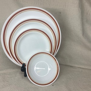 Puede incluir: Un juego de platos y un cuenco de cerámica blanca con bandas marrones y beige. Los platos son de diferentes tamaños, el más grande tiene un diámetro de unos 25 cm. El cuenco es más pequeño, con un diámetro de unos 15 cm.