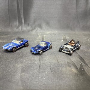 Puede incluir: Tres coches de juguete antiguos en tonos azules y plateados están dispuestos sobre una superficie oscura. Los coches incluyen un deportivo con techo negro, un vehículo de aspecto futurista y un modelo clásico descapotable. Los juguetes muestran signos de desgaste.