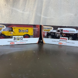 May include: Two Matchbox Collectibles die-cast model cars in their original boxes. The first car is a yellow 1925 Ford Model T delivery truck with a red roof and "Champion" branding. The second car is a black 1940 Ford delivery truck with a white roof and "Holley Performance" branding.
