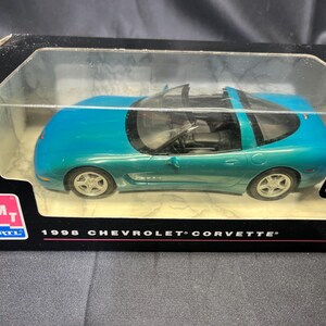 Puede incluir: Un coche modelo Chevrolet Corvette de 1998 de color turquesa en una vitrina de pl&aacute;stico transparente. El coche tiene ruedas plateadas y un interior negro. La caja tiene el logotipo de Chevrolet y el texto "1998 Chevrolet Corvette".