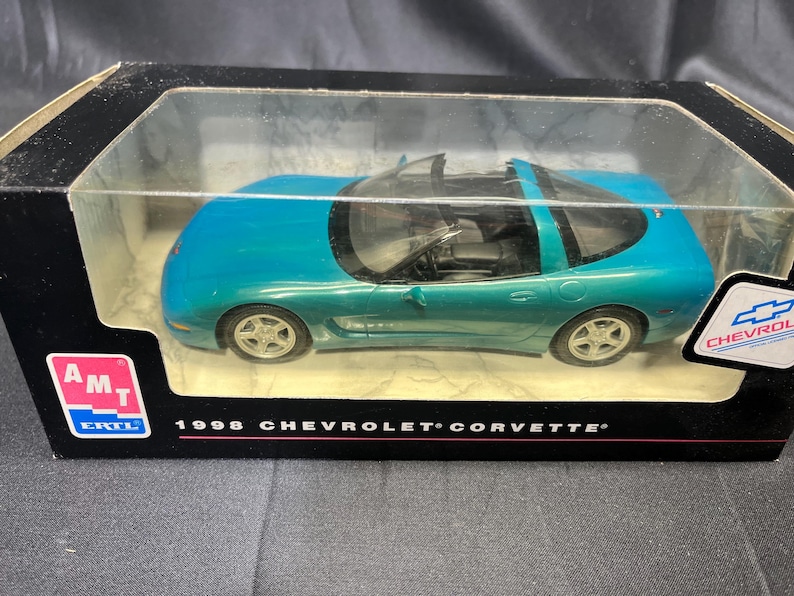 Puede incluir: Un coche modelo Chevrolet Corvette de 1998 de color turquesa en una vitrina de pl&aacute;stico transparente. El coche tiene ruedas plateadas y un interior negro. La caja tiene el logotipo de Chevrolet y el texto "1998 Chevrolet Corvette".