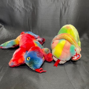 Puede incluir: Dos juguetes de peluche con forma de serpiente en colores vibrantes. Uno es principalmente rojo, azul y amarillo, mientras que el otro presenta un arcoíris de tonos. Ambos tienen lenguas rojas y ojos pequeños y redondos. Uno tiene una etiqueta.