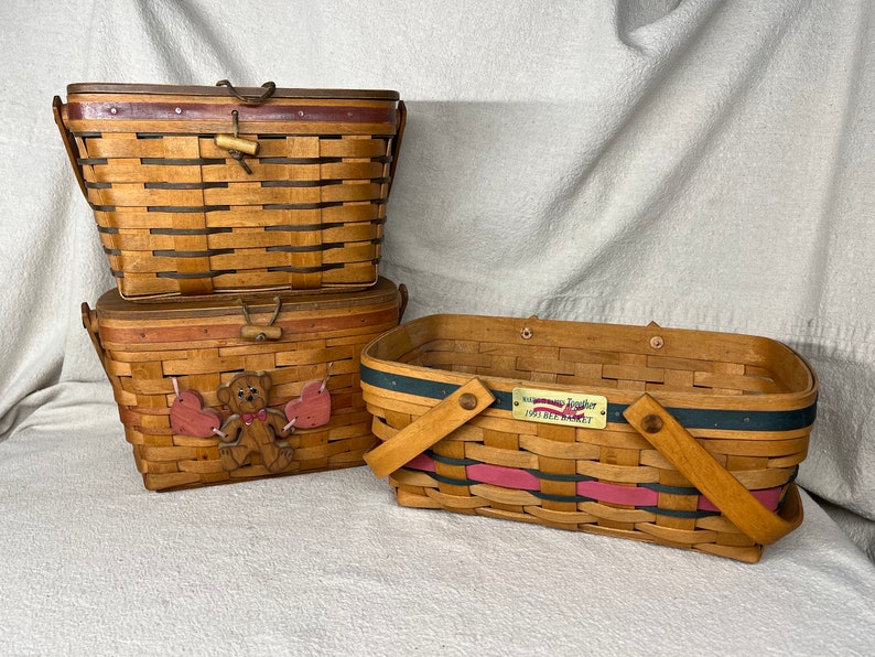 May include: Three wooden baskets with handles. The top basket is brown with a wooden latch. The middle basket is brown with a red and pink trim and a wooden bear with hearts. The bottom basket is brown with a blue and pink trim and a gold plaque that reads "Memories Together 1997 Bee Realty".