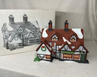 May include: A ceramic Christmas village building with a brown tiled roof, white timber framing, and snow detailing. The building has multiple windows and a sign that reads "Raspberry Inn". A monochrome drawing of the building is in the background.