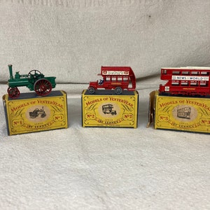 Puede incluir: Tres cajas de modelos de juguetes vintage de la serie Models of Yesteryear de Lesney. Las cajas son amarillas con letras rojas y negras. Las cajas presentan un autobús de dos pisos rojo, una máquina de vapor verde y un autobús de dos pisos rojo con la inscripción "NEWS WORLD" en el lateral.