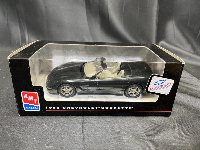 Puede incluir: Un modelo de coche negro de un Chevrolet Corvette de 1998 en una caja de presentaci&oacute;n. El modelo tiene un interior beige y ruedas plateadas. La caja tiene el logotipo de Chevrolet y el texto "1998 Chevrolet Corvette".