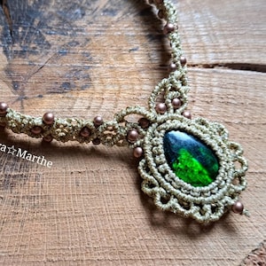 Può includere: Una collana in macramè dorata con un ciondolo di pietra preziosa verde e perline marroni. La collana è su una superficie di legno. Macra*Marthe è scritto sulla superficie.
