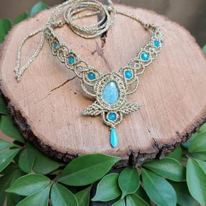 May include: A handmade macrame necklace with a central light blue stone and turquoise beads. The necklace is made with gold-colored cord and is displayed on a wooden surface with green leaves.