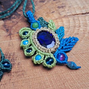 Può includere: Una collana macramè fatta a mano con un grande ciondolo ovale di pietra preziosa blu scuro con un bordo color oro. Il ciondolo è circondato da dettagli in macramè blu e verde, con perline blu e verdi aggiuntive. La collana è su un cordino verde.