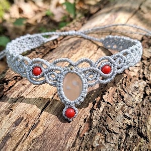 Può includere: Una collana girocollo in macramè grigio chiaro, con una pietra ovale centrale e perline rosse. La collana è esposta su un pezzo di legno, mettendo in risalto l'intricato nodo. Un delicato gioiello fatto a mano.