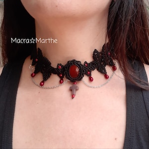 Può includere: Collana choker nera con una pietra rossa e una catena d'argento con un ciondolo a forma di croce. La collana è realizzata in macramè e ha perline rosse che pendono da essa. Il testo "Macra Marthe" è visibile nell'immagine.