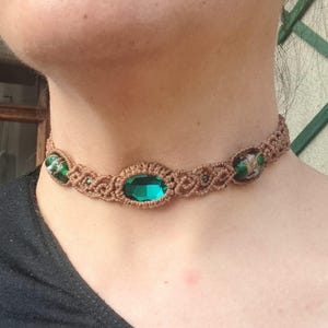 Op de afbeelding: Een bruine macramé choker ketting met groene glazen kralen en een grote smaragdgroene edelsteen.