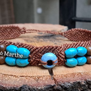 May include: A brown macrame bracelet with turquoise and wood beads. The bracelet has a blue and black eye bead in the center. The bracelet is on a wooden surface. Macra*Marthe is written on the bracelet.