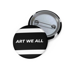 Op de afbeelding: Twee ronde zwart-witte buttons. De voorste button heeft de tekst "ART WE ALL" in het wit. De button heeft een zwarte achtergrond met twee witte horizontale strepen. De tweede button ligt met de achterkant naar boven.