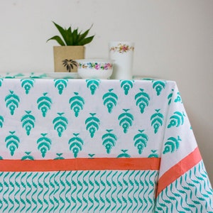 May include: White tablecloth with a teal leaf pattern and a coral border. The tablecloth is decorated with a repeating teal leaf design and a coral border with a teal wavy pattern. A small plant and dishware are visible in the background.