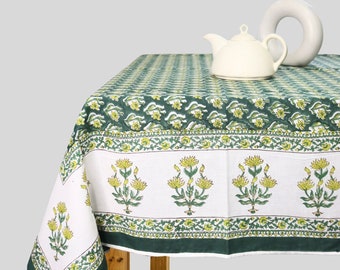 May include: A green and white tablecloth with a floral pattern. The tablecloth is laid on a wooden table with a white teapot and a white vase with a dried branch.