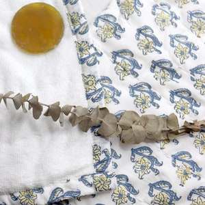 May include: A white cotton towel with a blue and yellow floral print border. A round, amber-colored soap bar is on the towel. A sprig of dried eucalyptus is on the towel.