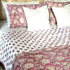 May include: A bed with a white sheet, a patterned quilt, and pillows. The quilt features a repeating pattern of brown floral shapes on a white background, transitioning to a larger floral design in red and beige. White, textured pillows and patterned pillowcases complete the bedding.