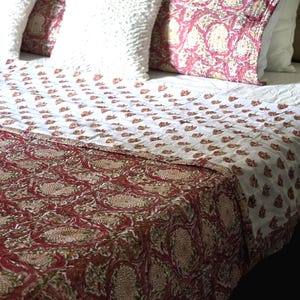 May include: A bed is made with a white and red floral patterned quilt and matching decorative pillows. The pillows are white and red with a floral design. The bed has a white headboard and white sheets.