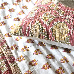 May include: A close-up of a bed set featuring a white quilt and pillow with a floral pattern in pink, yellow, and green. The quilt has a repeating pattern of leaves, while the pillow has a larger, more detailed floral design.