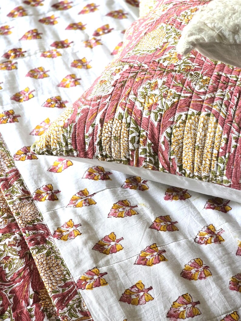 May include: A close-up of a bed set featuring a white quilt and pillow with a floral pattern in pink, yellow, and green. The quilt has a repeating pattern of leaves, while the pillow has a larger, more detailed floral design.