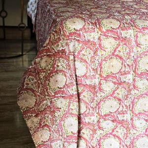 May include: A floral patterned quilt in shades of pink, cream, and green. The quilt features a repeating design of stylized flowers and leaves. The bedding is draped over a bed, with a matching pillow visible. The overall theme is home decor.