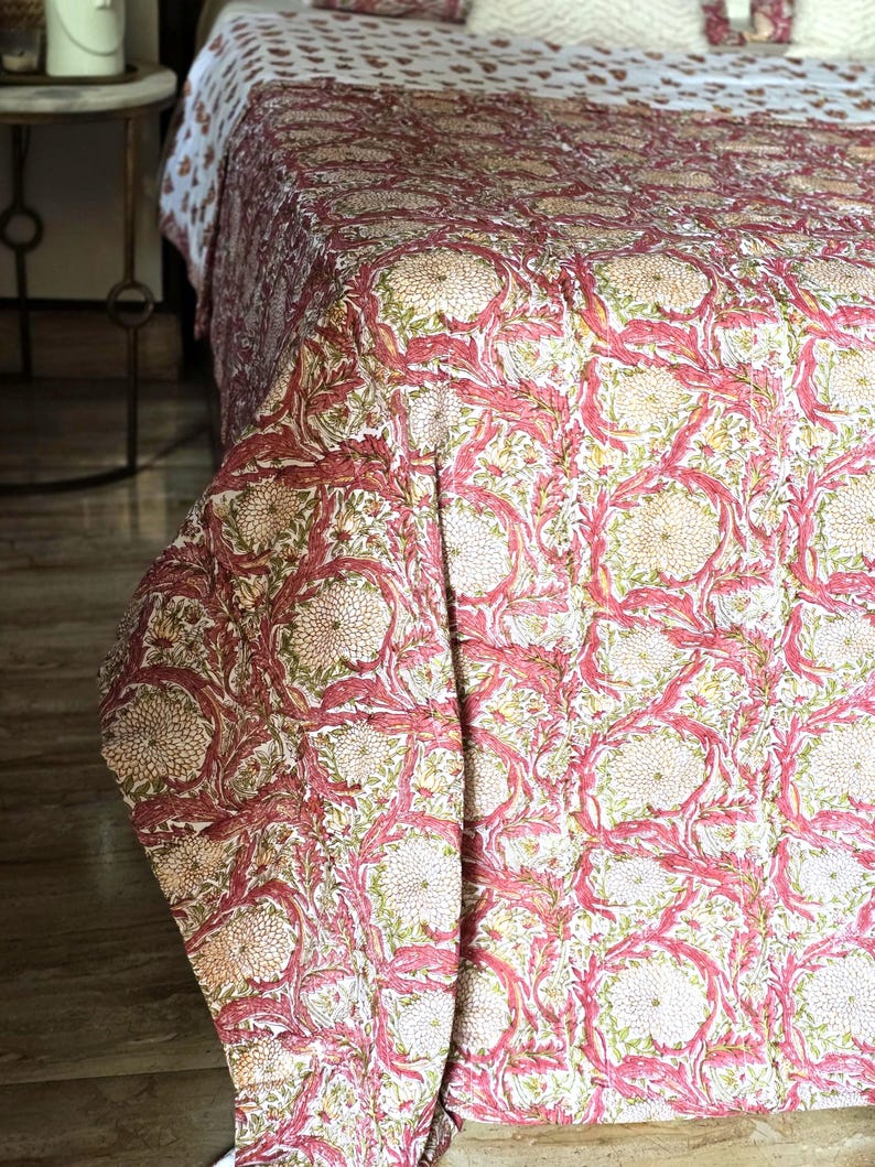 May include: A floral patterned quilt in shades of pink, cream, and green. The quilt features a repeating design of stylized flowers and leaves. The bedding is draped over a bed, with a matching pillow visible. The overall theme is home decor.
