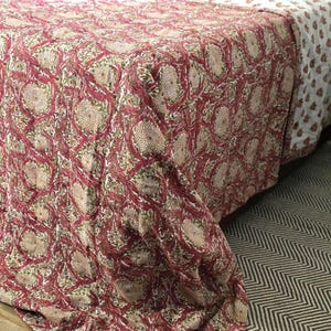 May include: A bedspread with a floral pattern in shades of red, beige, and green. The design features large, stylized flowers and intertwining vines. The bedspread is draped over a bed, with a white sheet and patterned pillows visible.